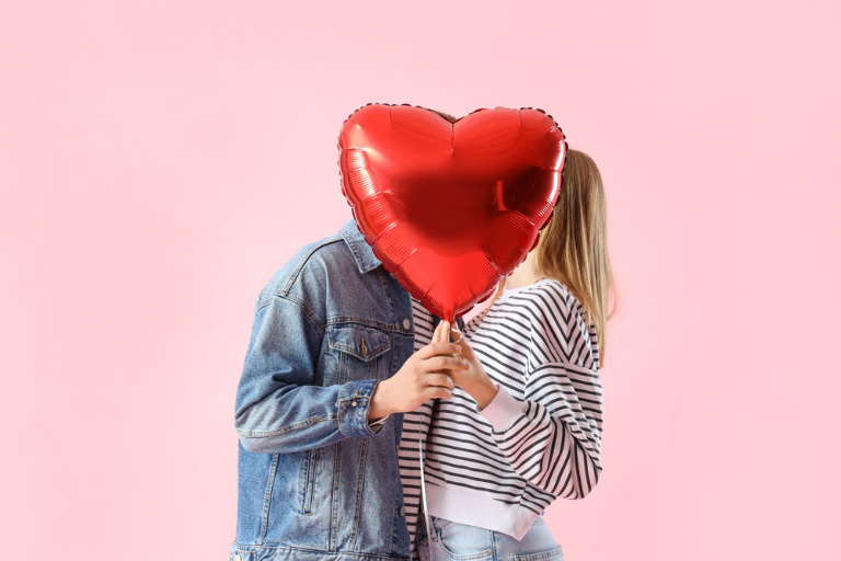 Happy young couple with heart-shaped balloon on pink background.